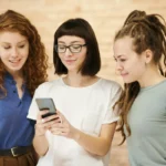 Tiga perempuan tersenyum bersama melihat layar ponsel, menggambarkan suasana berbagi rekomendasi dalam program referral